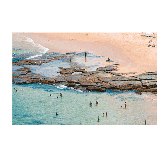 A close-up aerial view of Bateau Bay, NSW, capturing swimmers enjoying the shallow, crystal-clear waters near the rocky shoreline. The warm golden sand contrasts with the turquoise sea, creating a vibrant coastal scene.