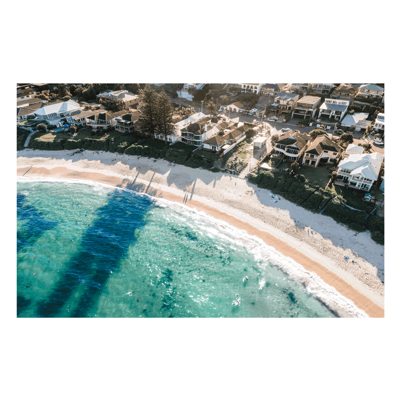 Aerial view of Blue Bay, NSW, featuring turquoise waters, golden sand, and beachfront homes with long afternoon shadows cast by Norfolk pines.