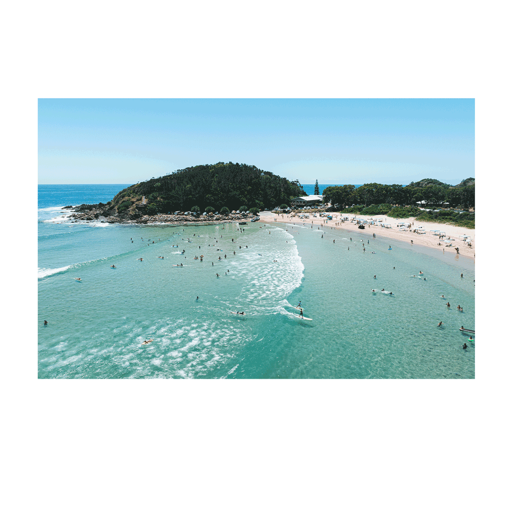 A stunning aerial view of Scotts Head, NSW, showing crystal-clear turquoise waters, surfers riding gentle waves, and a bustling beach filled with holidaymakers. The lush green headland frames the picturesque coastal scene under a vibrant blue sky.