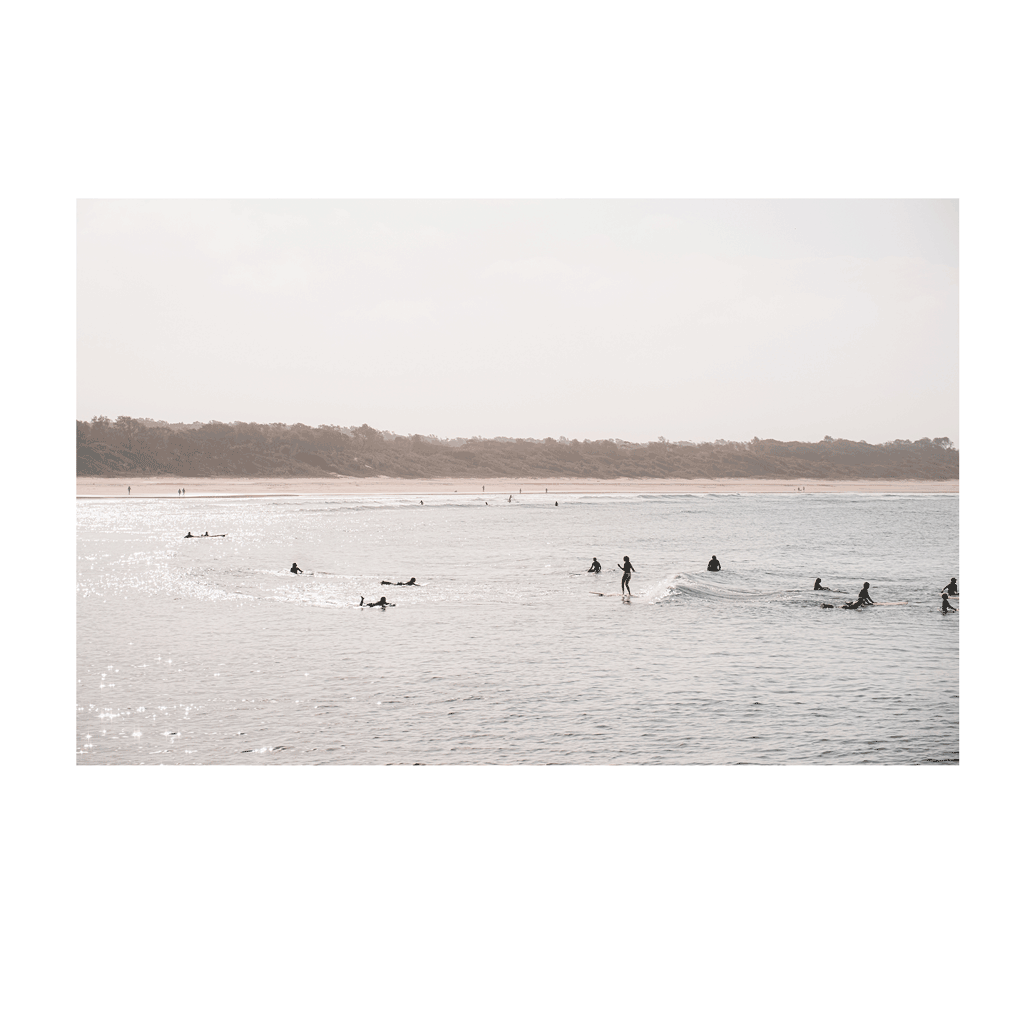 A serene sunset surf scene at Scotts Head, NSW, with surfers gliding across the shimmering ocean as golden light reflects off the water. The distant shoreline and soft pastel sky create a peaceful coastal moment.