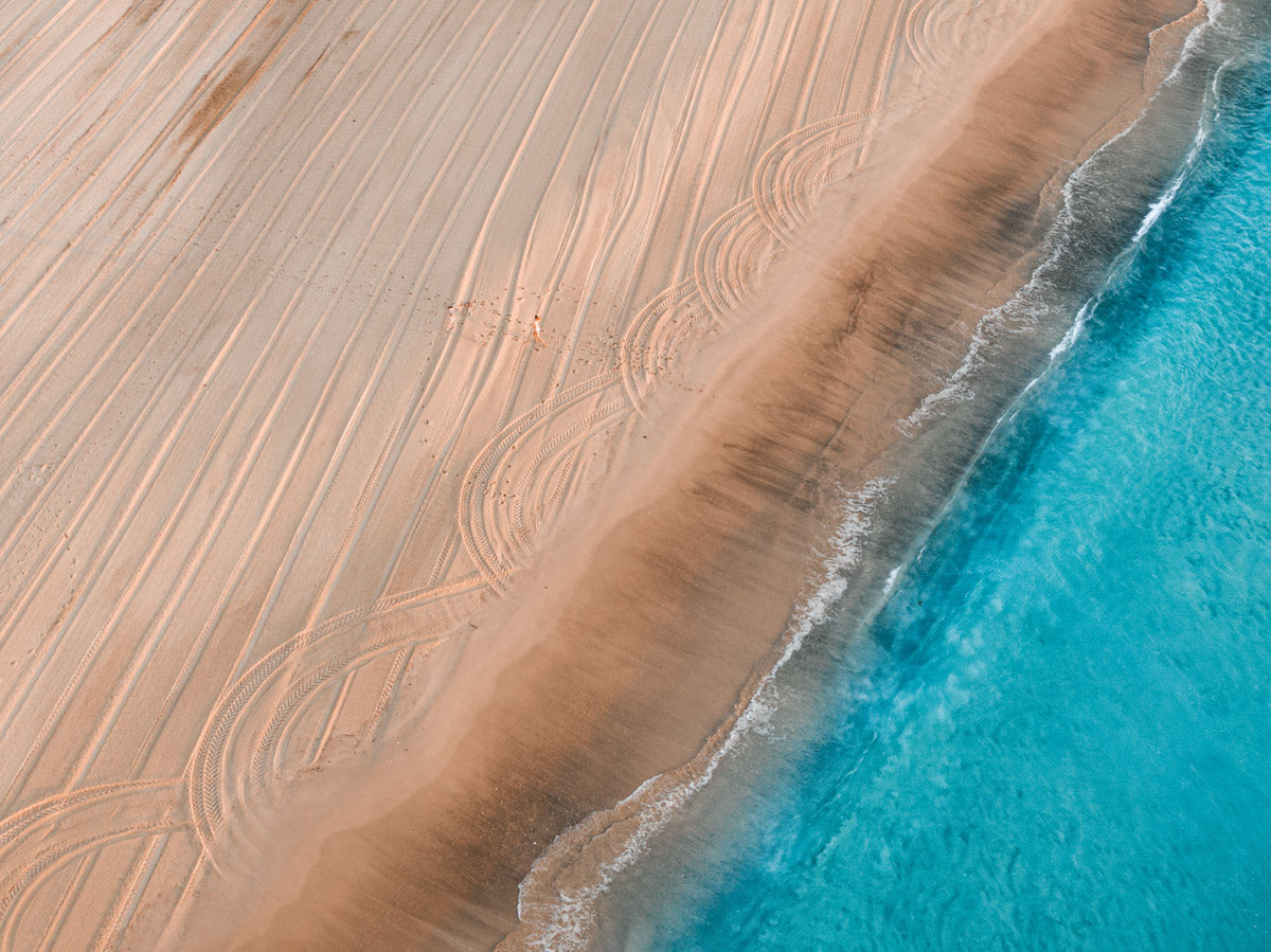 Aerial view of Catherine Hill Bay’s sandy beach, featuring sweeping tyre tracks and vibrant turquoise waves meeting the shore.