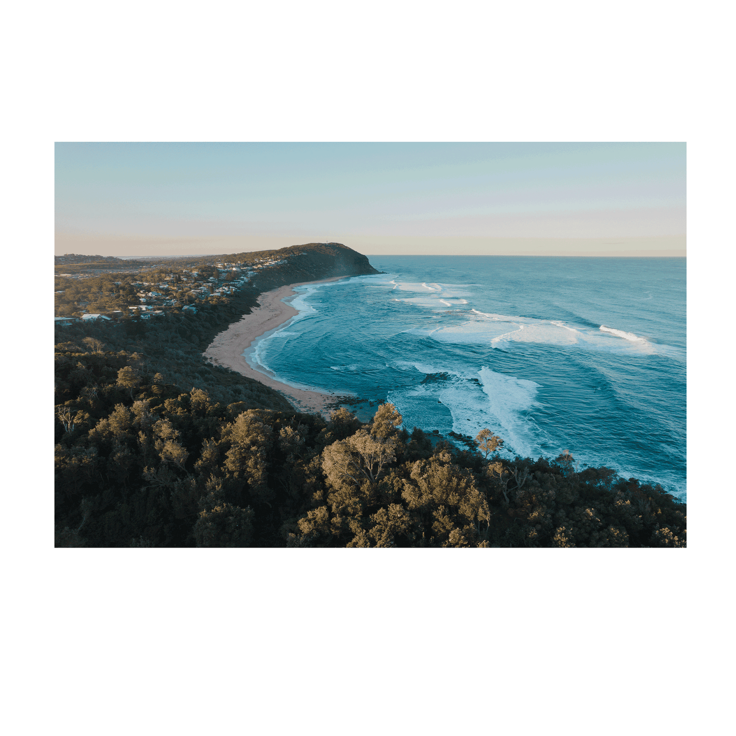 Alt Text: A breathtaking aerial view of Forresters Beach, NSW, featuring a rugged headland, rolling waves, and golden sand under a soft morning light.
