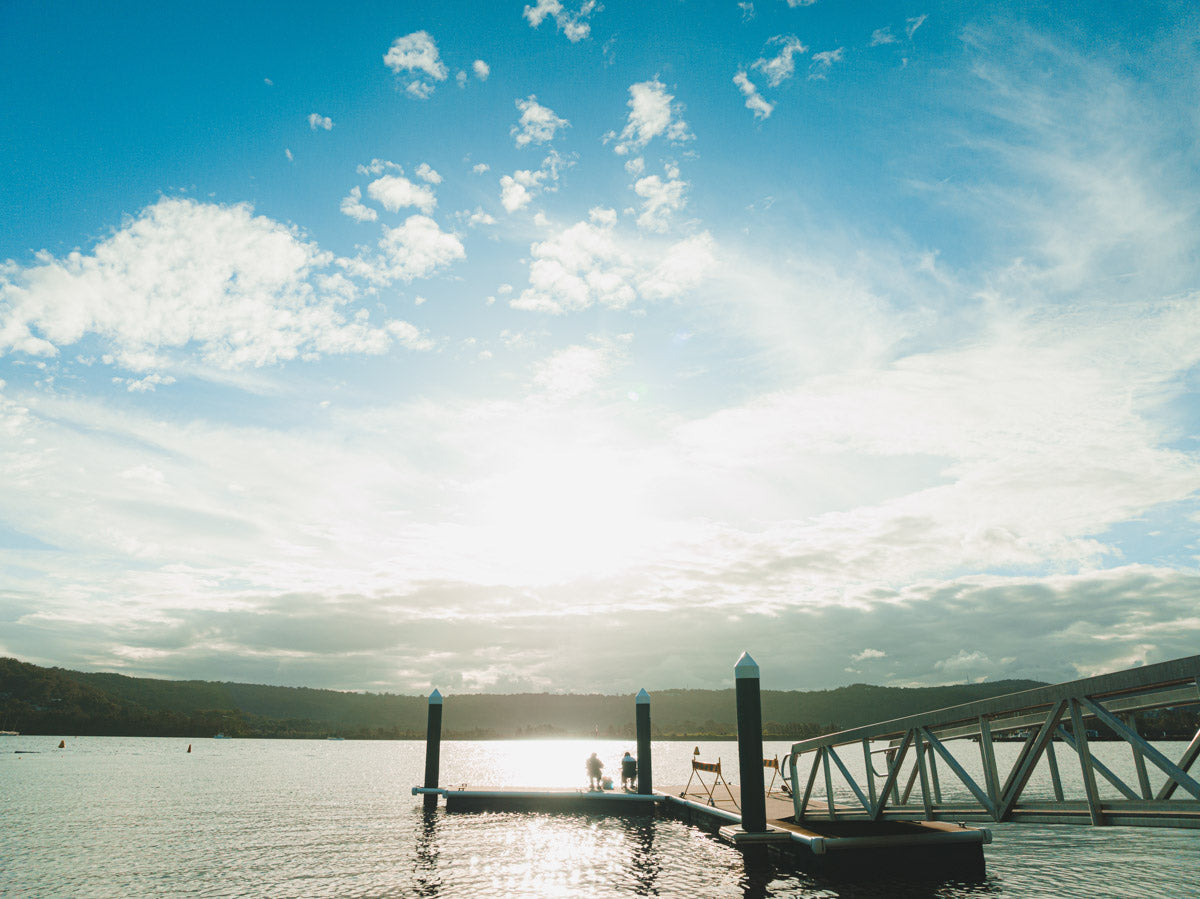 A tranquil scene at a jetty in Gosford, NSW, with two figures sitting by the water as the sun casts a golden reflection on the calm surface.