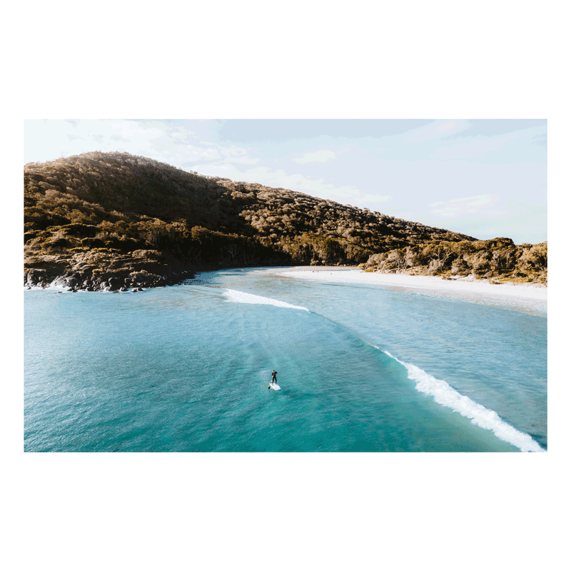 A stunning aerial view of Hat Head, NSW, featuring a lone surfer riding a wave against a backdrop of lush hills and pristine waters.