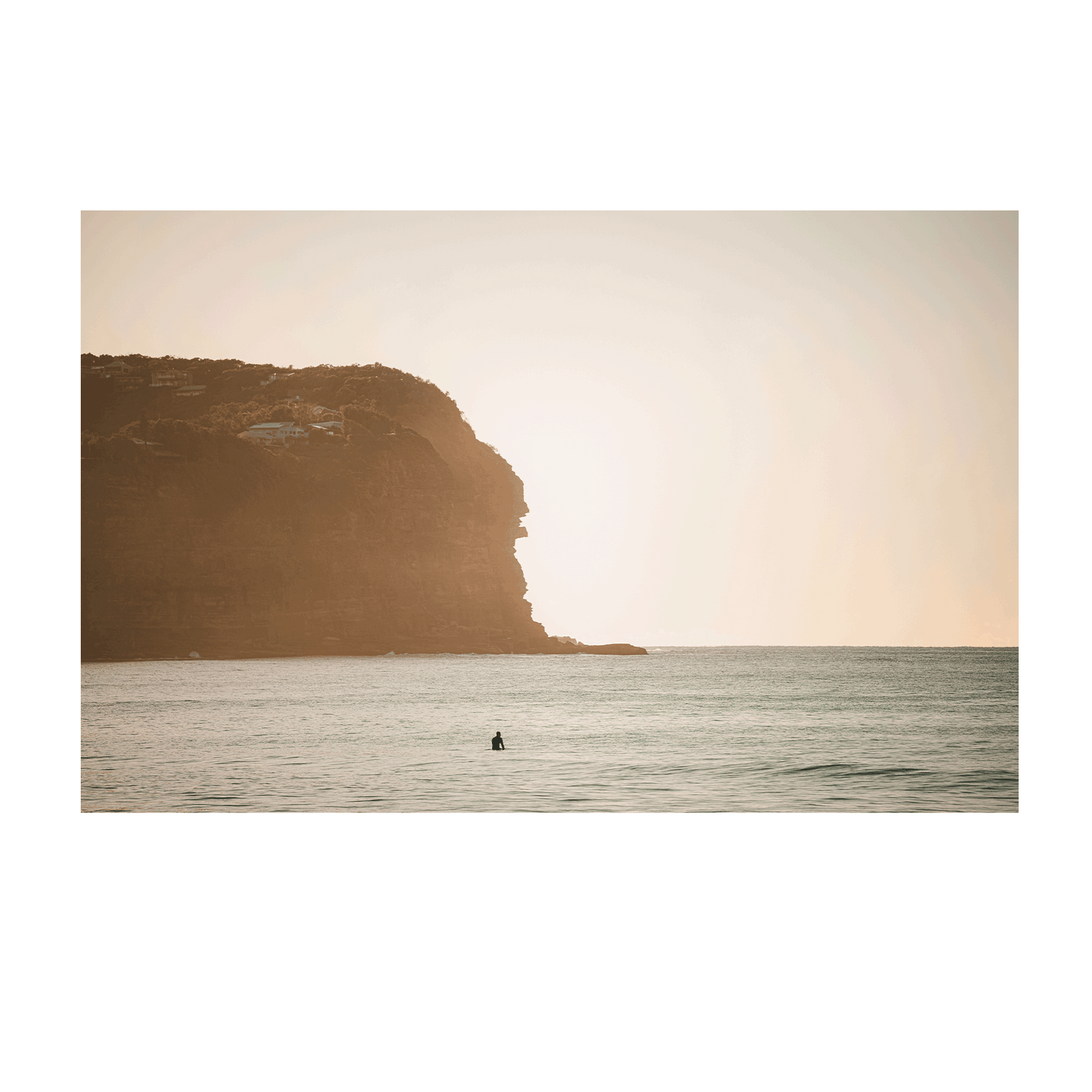 A lone surfer waits in the golden sunrise at Macmasters Beach, NSW, with a towering cliff bathed in soft morning light.