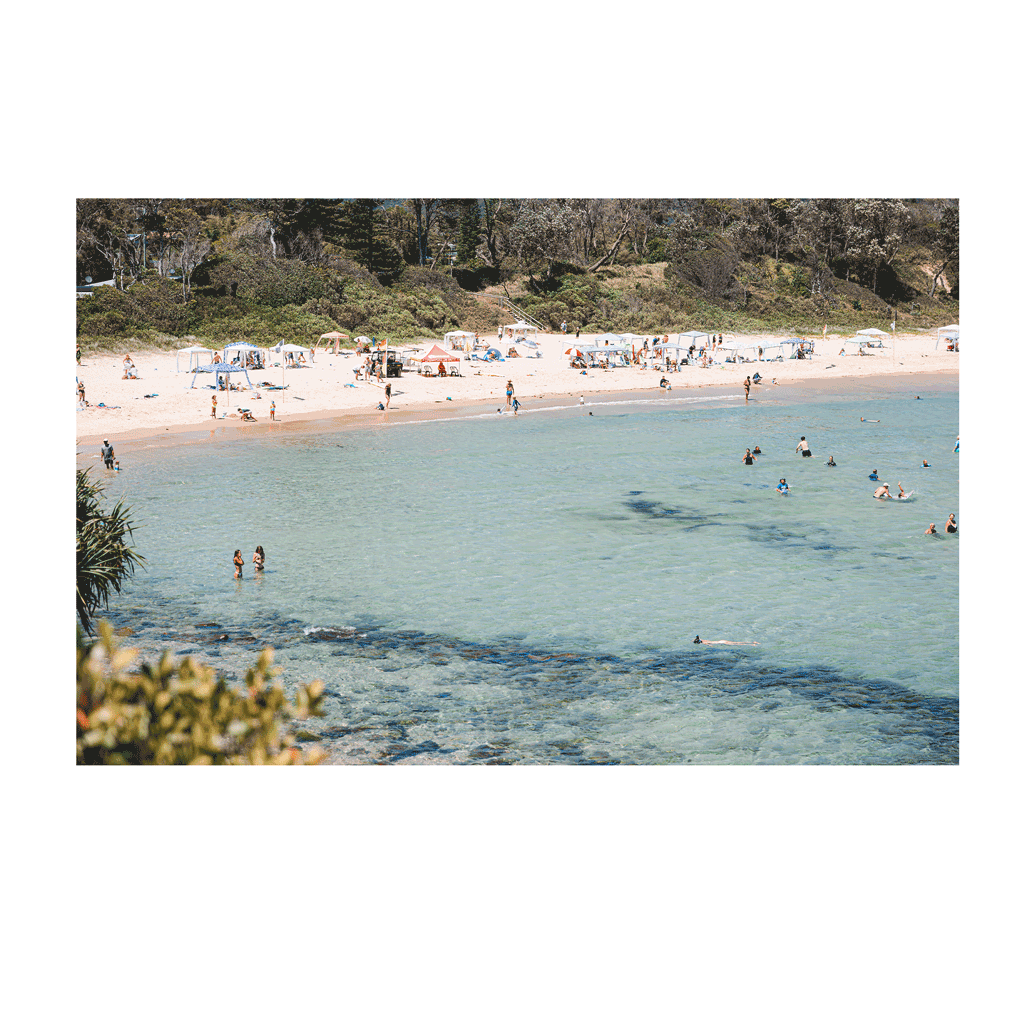 A lively summer scene at Scotts Head, NSW, featuring clear turquoise waters, swimmers enjoying the refreshing ocean, and sunbathers relaxing along the golden sandy beach. The vibrant coastal setting captures the essence of a perfect beach day.
