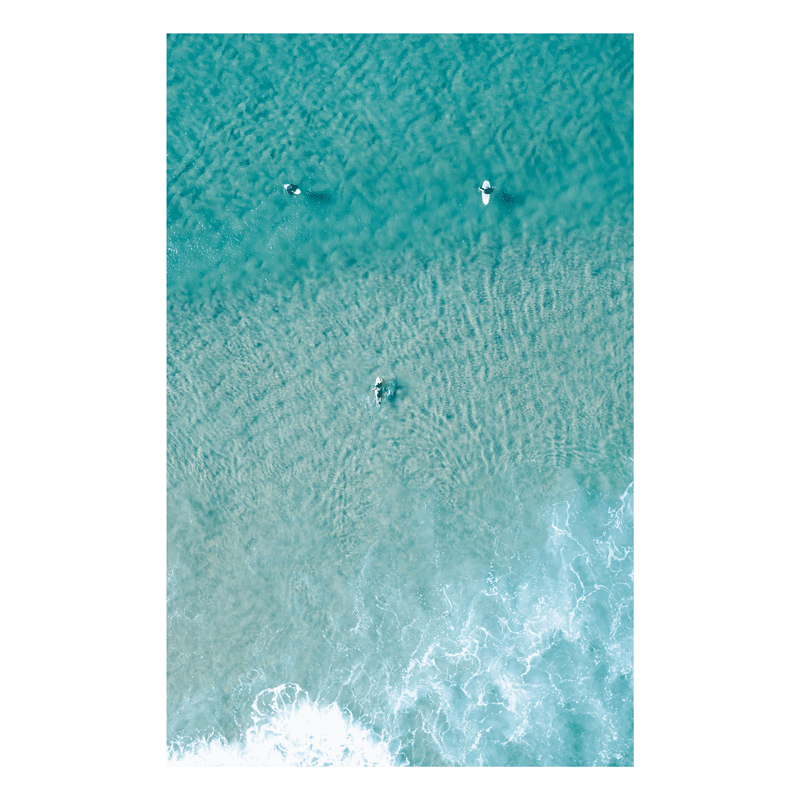 📸 An aerial view of Wamberal Beach, NSW, featuring three surfers in crystal-clear turquoise waters, with soft waves rolling onto the shore.