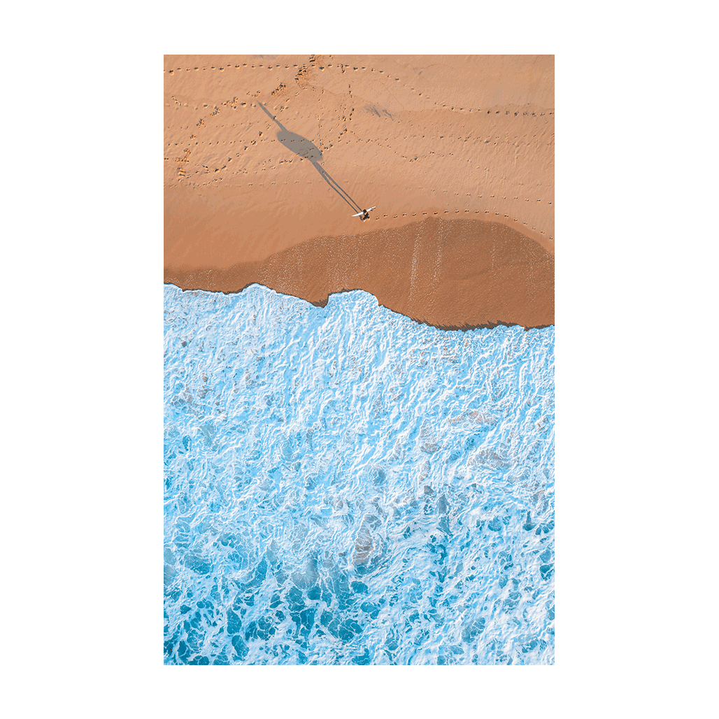 An aerial view of Wamberal Beach, NSW, featuring a lone surfer walking along golden sand, casting a long shadow as waves crash onto the shore.