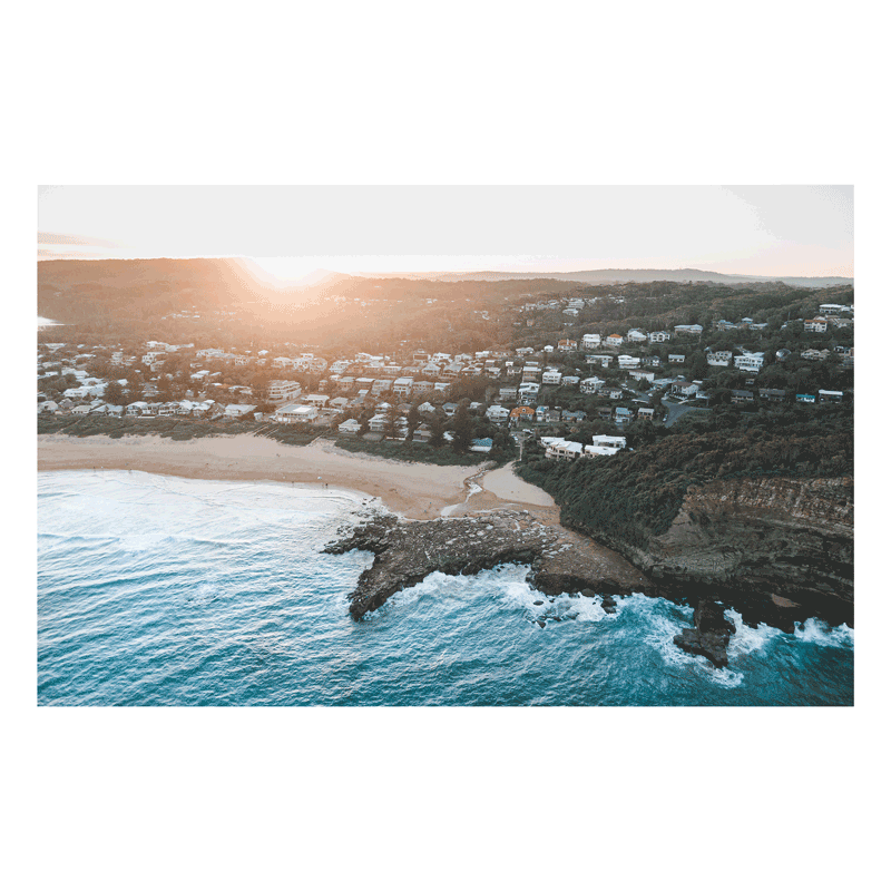 Aerial view of North Avoca Beach, NSW, at sunset, with golden light illuminating the coastal hillside, ocean waves, and rocky shoreline.