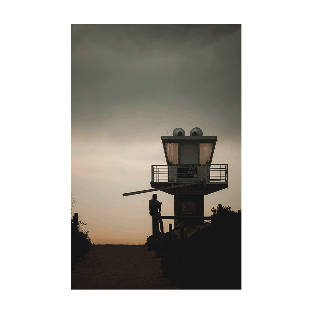 A moody coastal scene at Avoca Beach, NSW, featuring a silhouetted figure near a lifeguard tower under a dramatic sky at dusk.