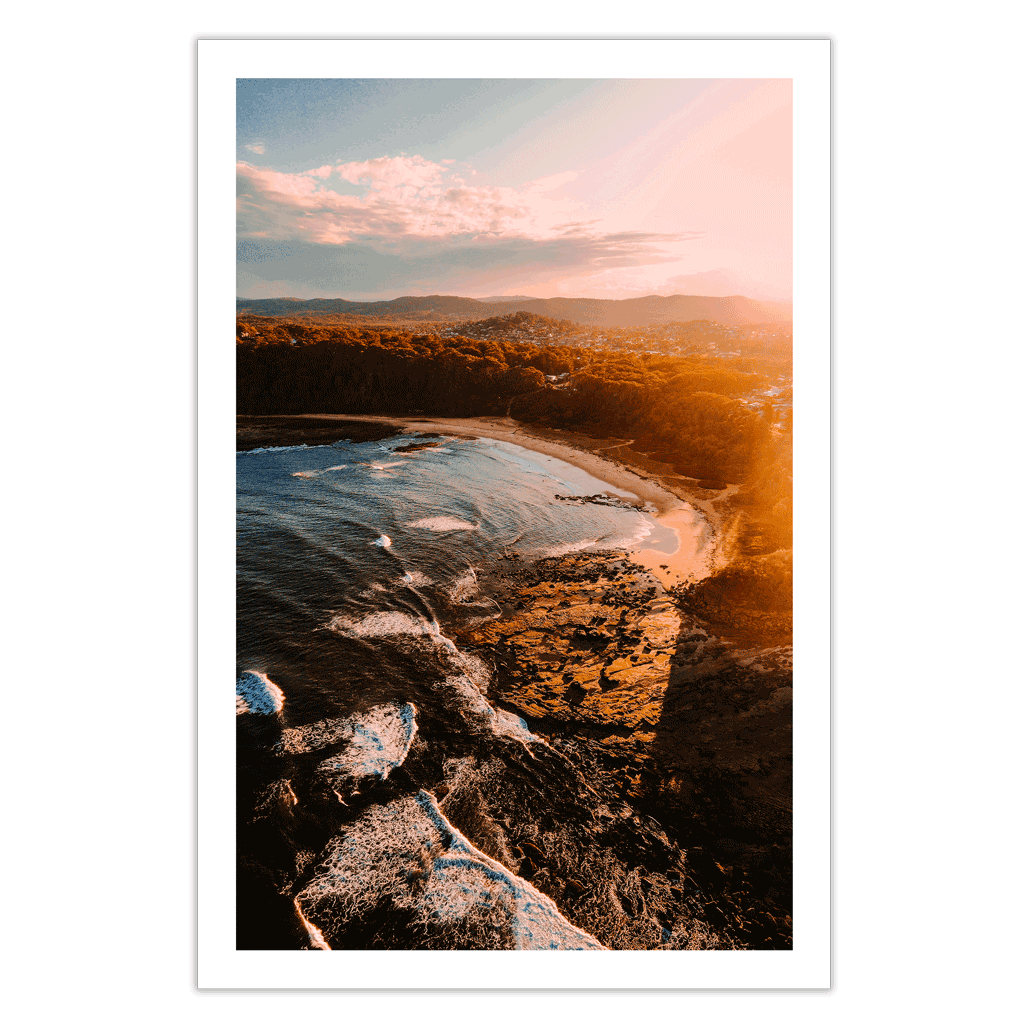 Aerial view of Bateau Bay, NSW, capturing golden light reflecting on the rugged coastline as waves crash against the shore at sunset.