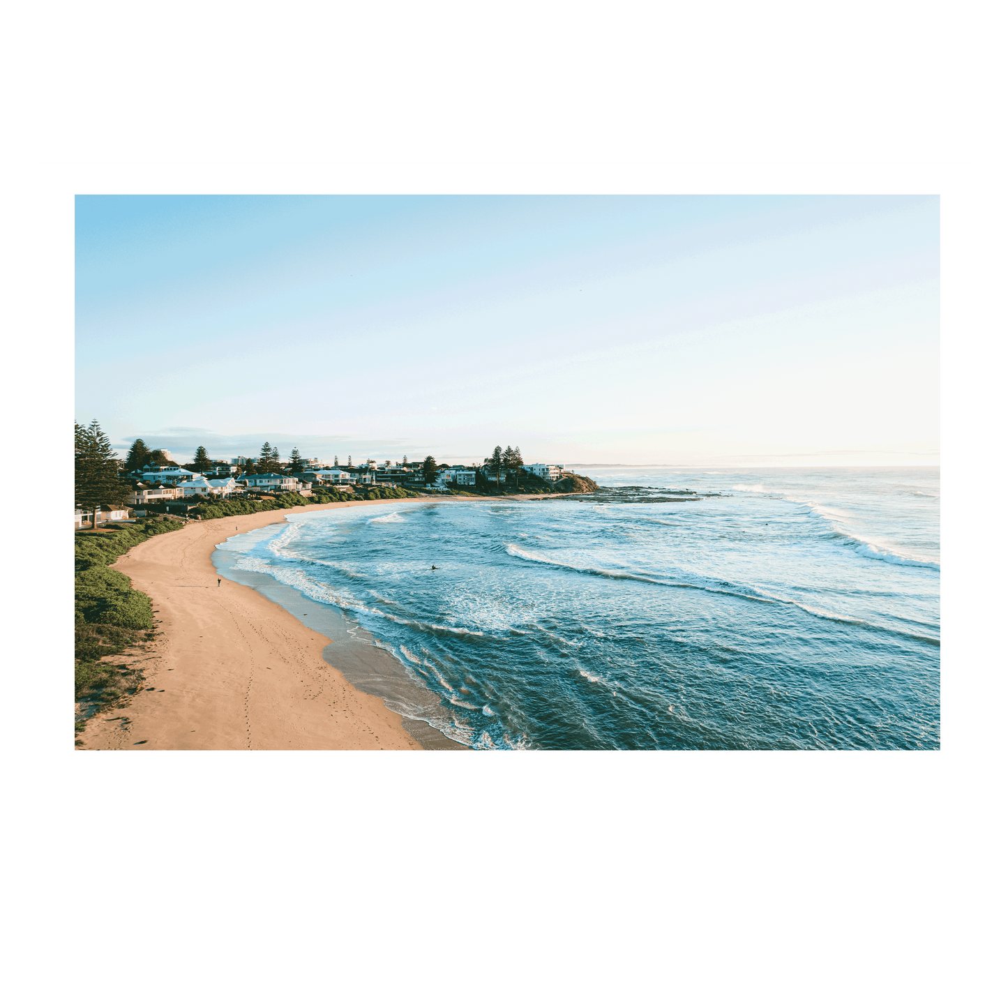A scenic morning view of Blue Bay, NSW, featuring rolling waves, golden sand, and coastal homes under soft winter light.