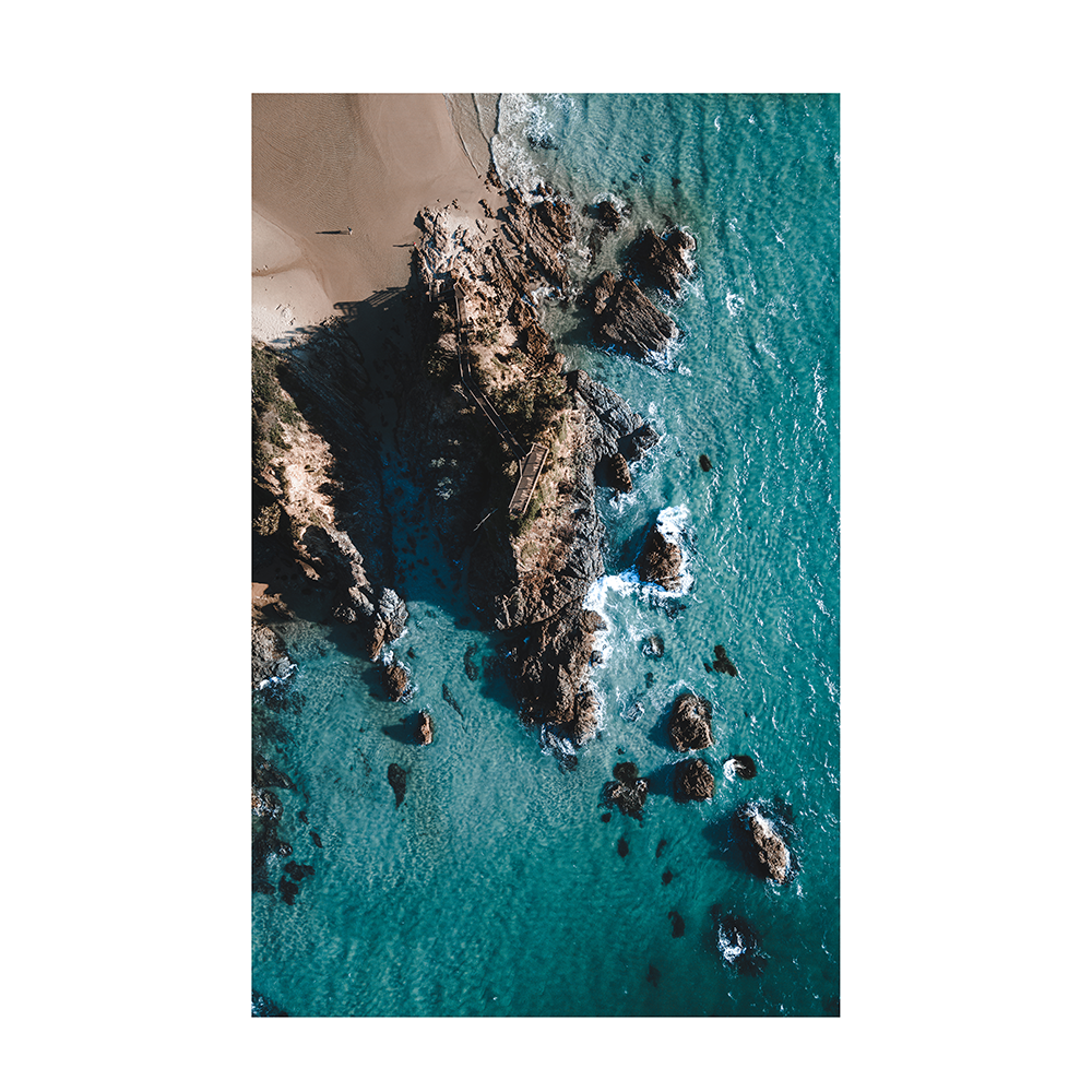An aerial view of The Pass, Byron Bay, NSW, showcasing turquoise waters, rocky outcrops, and a lone figure walking along the beach.
