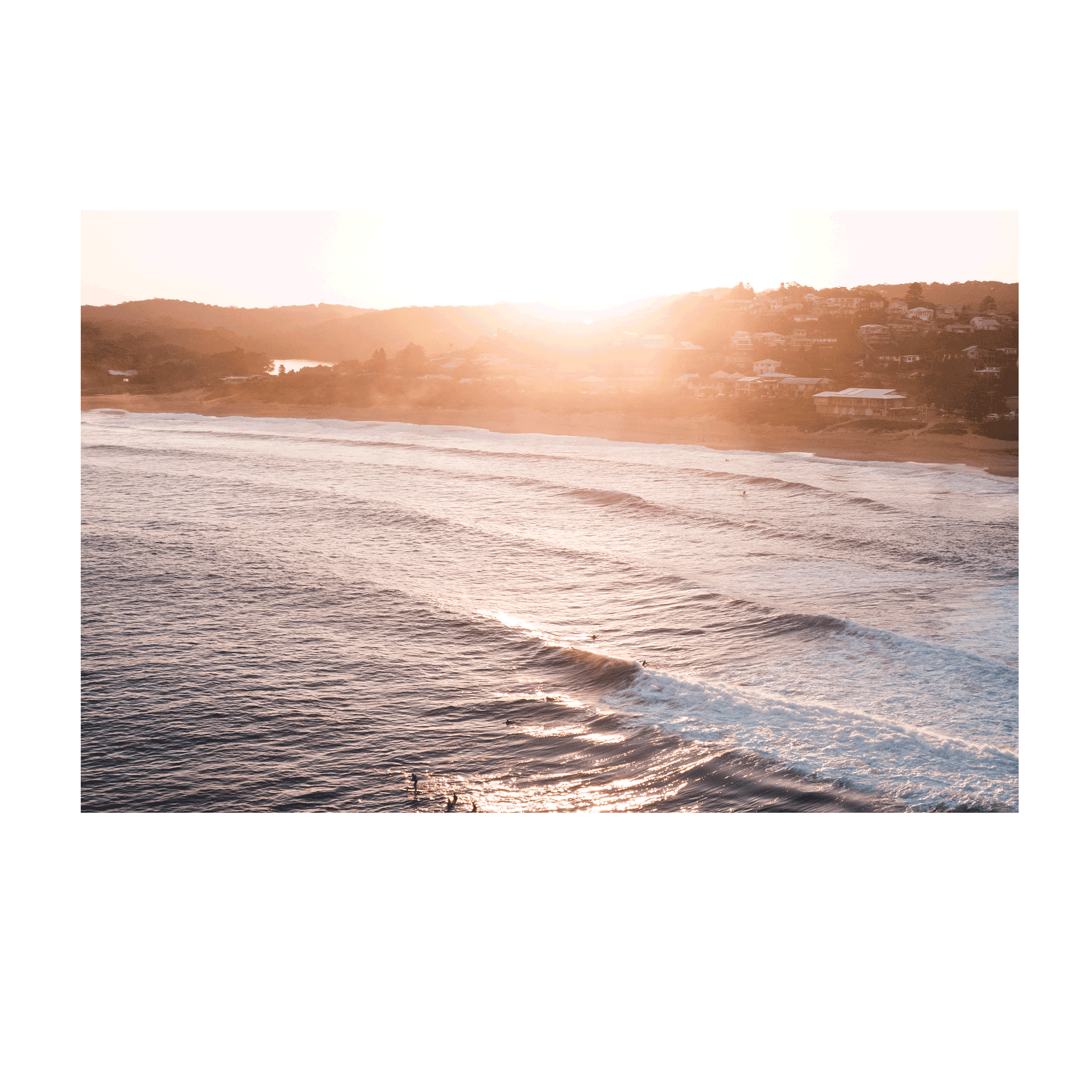 Aerial view of Copacabana Beach, NSW, at sunset, featuring rolling waves glowing under the setting sun with a warm coastal backdrop.
