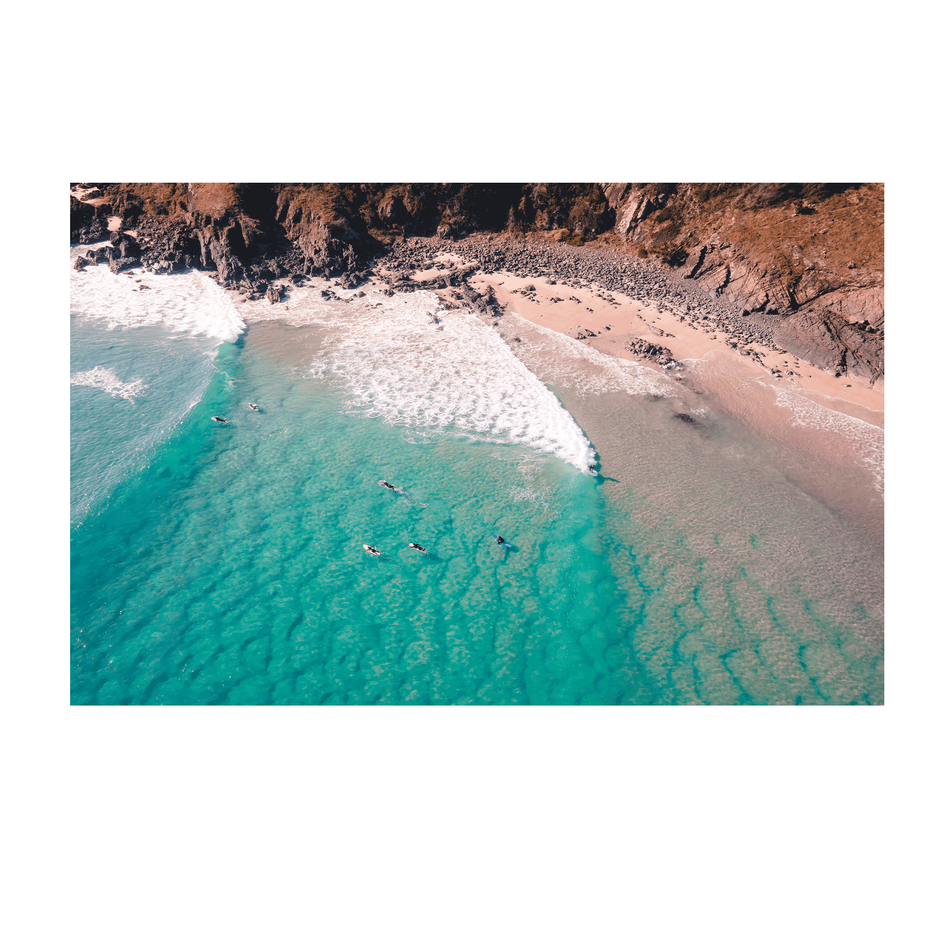 A stunning aerial view of Racecourse, Crescent Head, NSW, featuring surfers paddling in turquoise waves near a rugged coastline.
