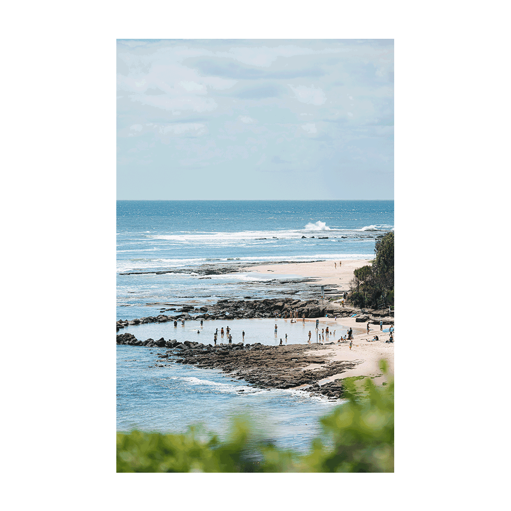 A scenic coastal view of Norah Head Beach, NSW, featuring natural rock pools filled with beachgoers, with waves rolling in the distance.