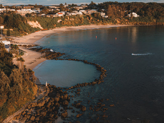 Aerial view of Norah Head, NSW, showcasing the ocean pool surrounded by rocks, sandy beach, and coastal homes in the soft afternoon light.