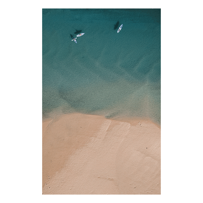 An aerial view of Red Rock, NSW, showcasing turquoise waters and sandy beaches, with two paddleboarders gliding across the bay.
