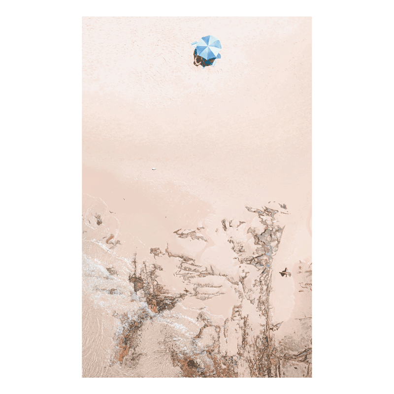 Experience the beauty of simplicity with Below – a minimalist aerial print of Spoon Bay, showcasing a lone blue umbrella on warm sands with textured rock formations.