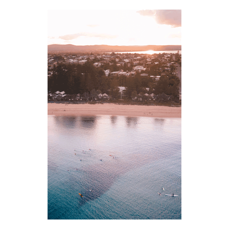 Aerial view of Toowoon Bay Beach at sunset, with golden light reflecting off calm waters. Paddleboarders and kayakers glide across the bay, creating a peaceful coastal scene perfect for a serene and modern space.
